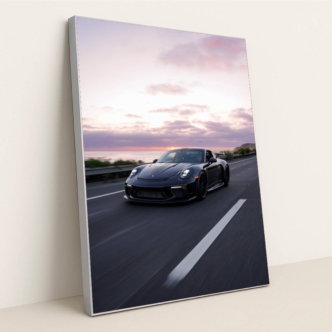 This photo shows a silver Porsche sports car driving on a winding mountain road at sunset, in a silver frame.
