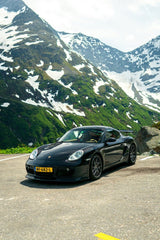 Black Porsche Cayman in the Alps, a sleek car on a mountain road with snow-capped peaks in the background for car wall art.