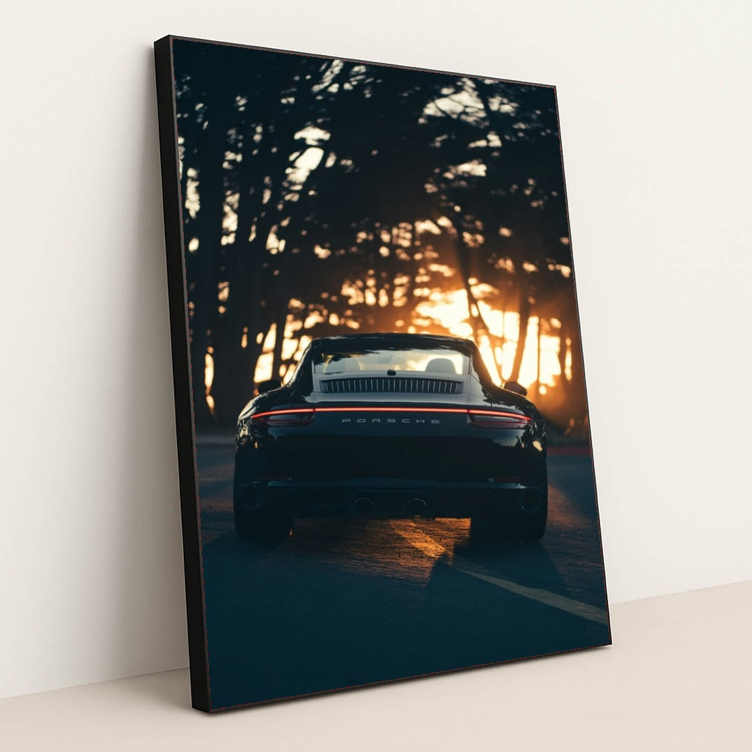 This photo shows the rear view of a black convertible Porsche parked on a tree-lined road at sunset, in a black frame.