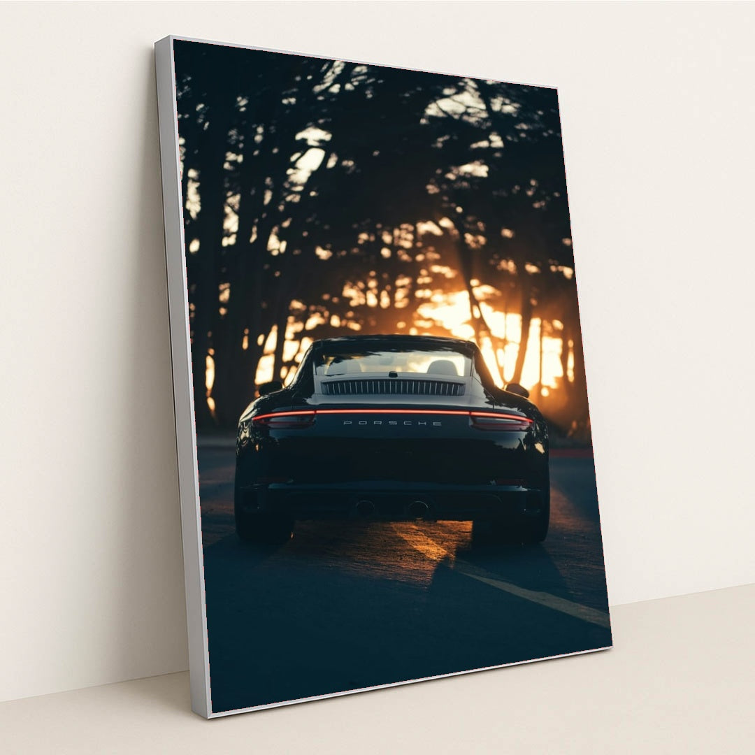 This photo shows the rear view of a black convertible Porsche parked on a tree-lined road at sunset, in a silver frame.