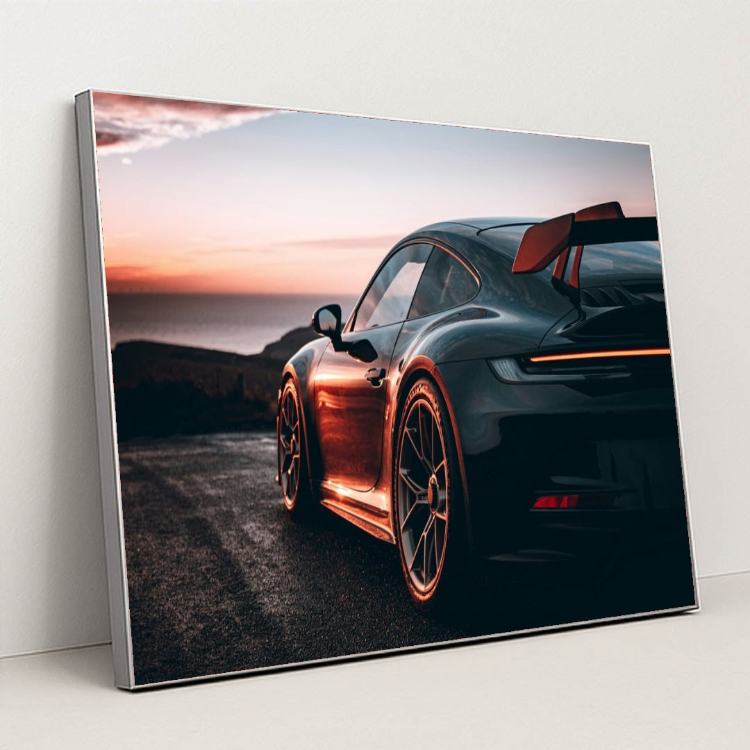 This photo shows a black Porsche sports car with a large rear wing parked at sunset near the ocean, in a silver frame.