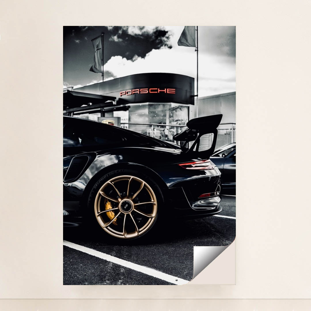 This photo shows the rear side view of a black Porsche GT sports car with gold wheels parked outside a dealership, minimalist unframed style.