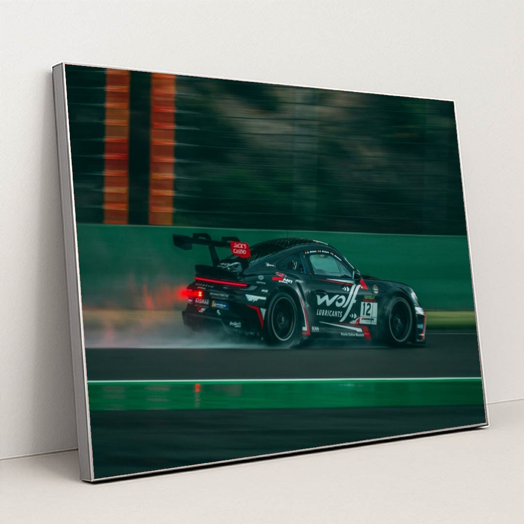 This photo shows a black Porsche race car with sponsor decals driving fast on a wet racetrack, in a silver frame.