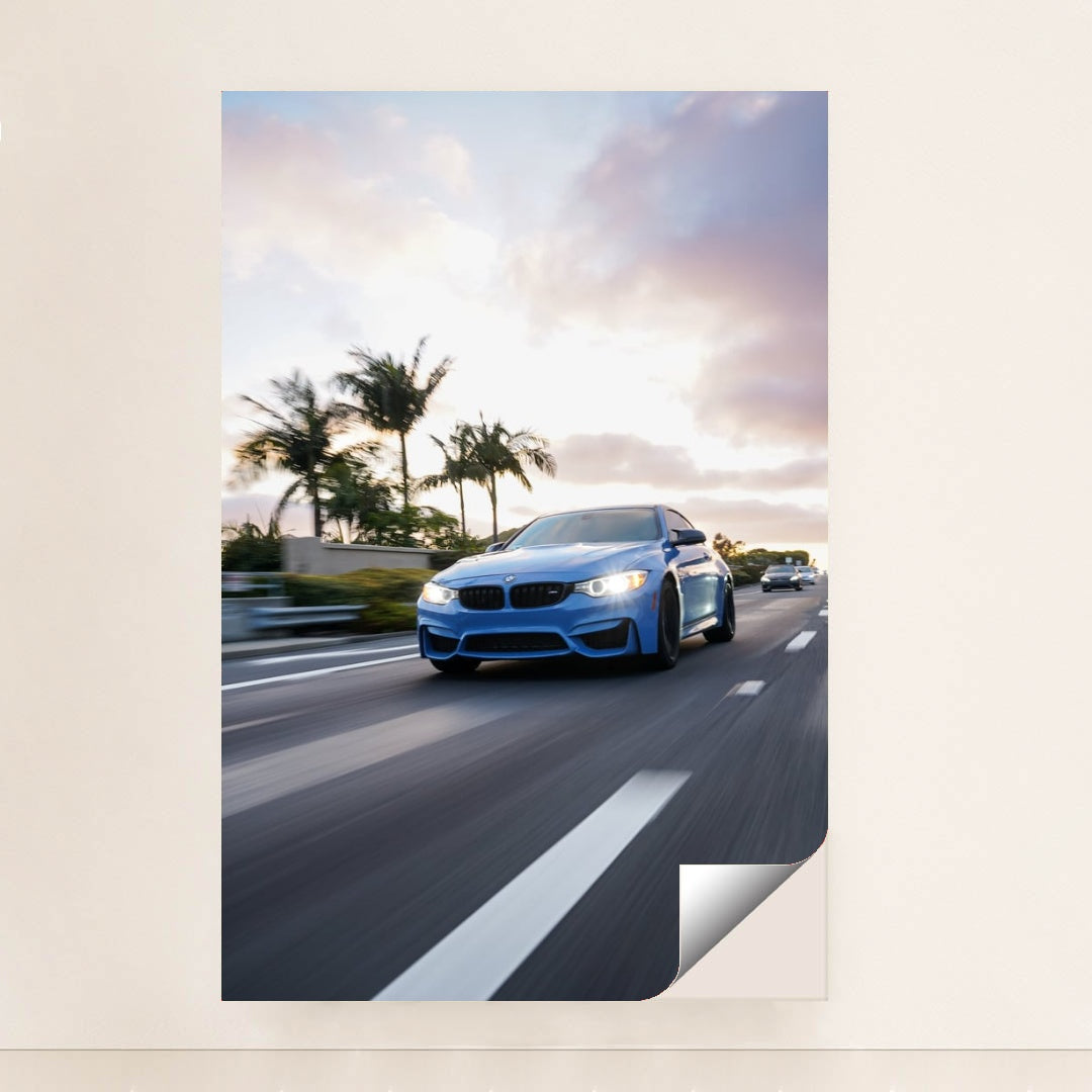 This photo shows a blue BMW driving down a palm-tree-lined highway at sunset, unframed.