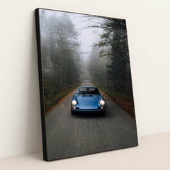 This photo shows a classic blue Porsche 911 parked on an empty forest road in morning mist, in a black frame.