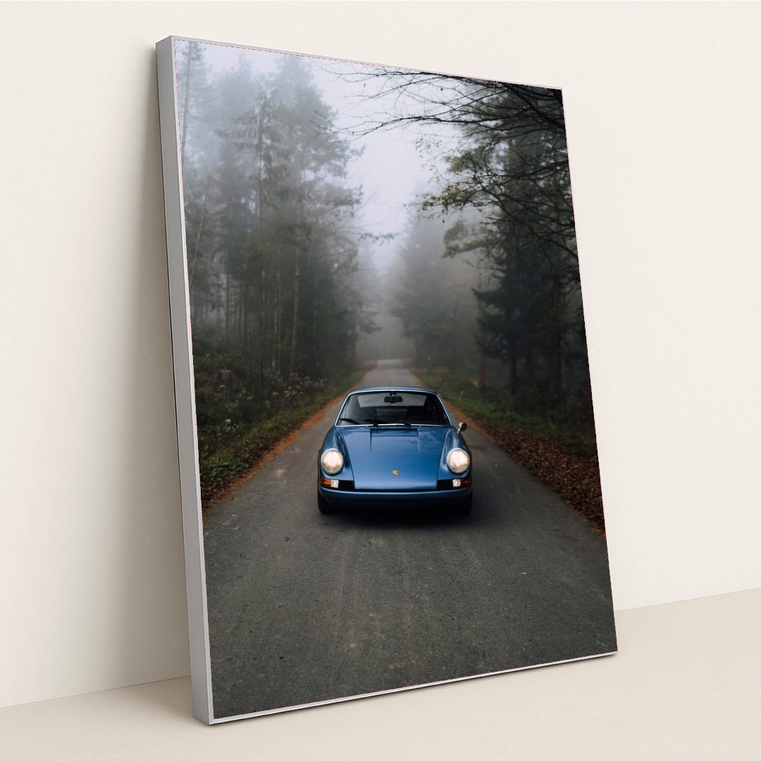 This photo shows a classic blue Porsche 911 parked on an empty forest road in morning mist, in a silver frame.