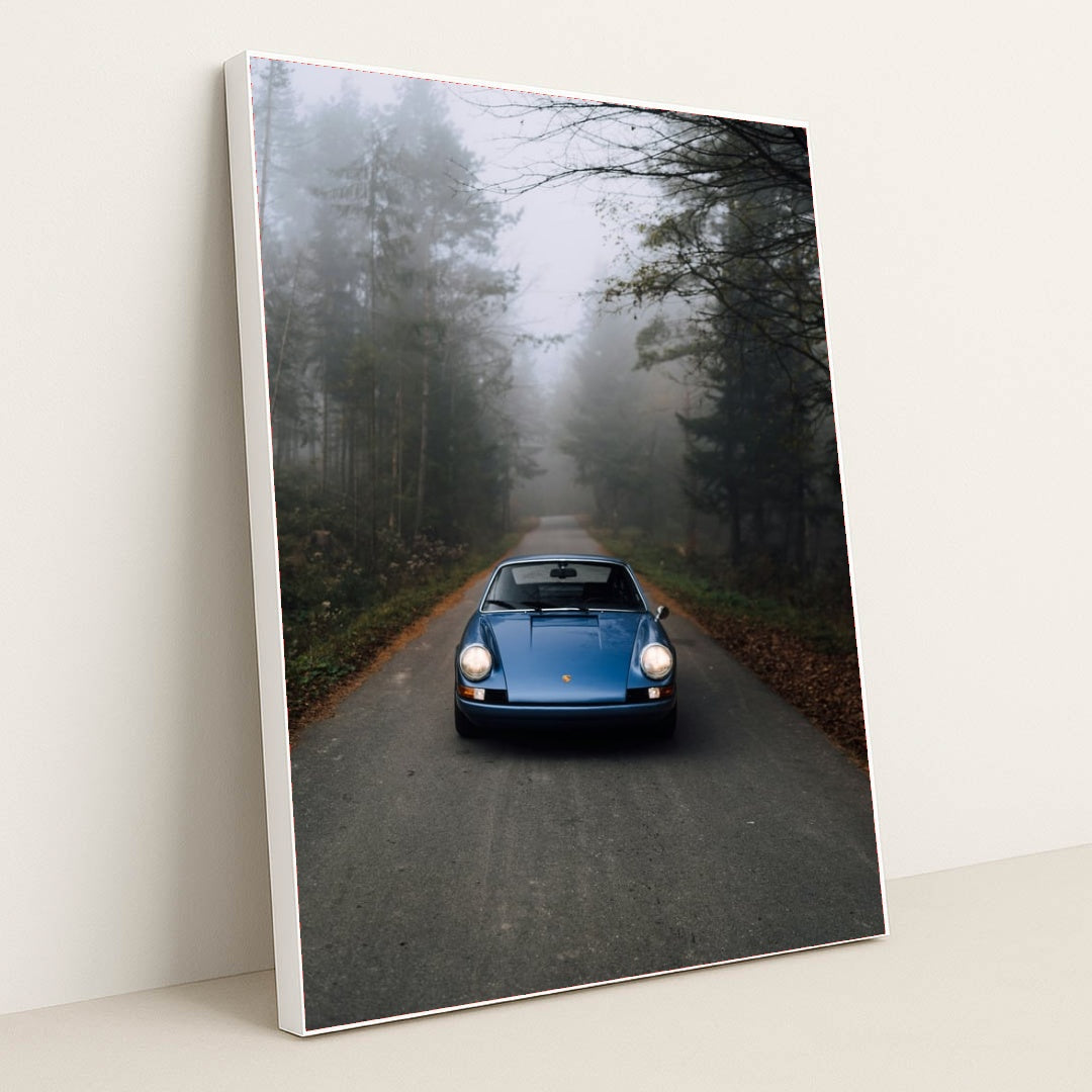 This photo shows a classic blue Porsche 911 parked on an empty forest road in morning mist, in a white frame.