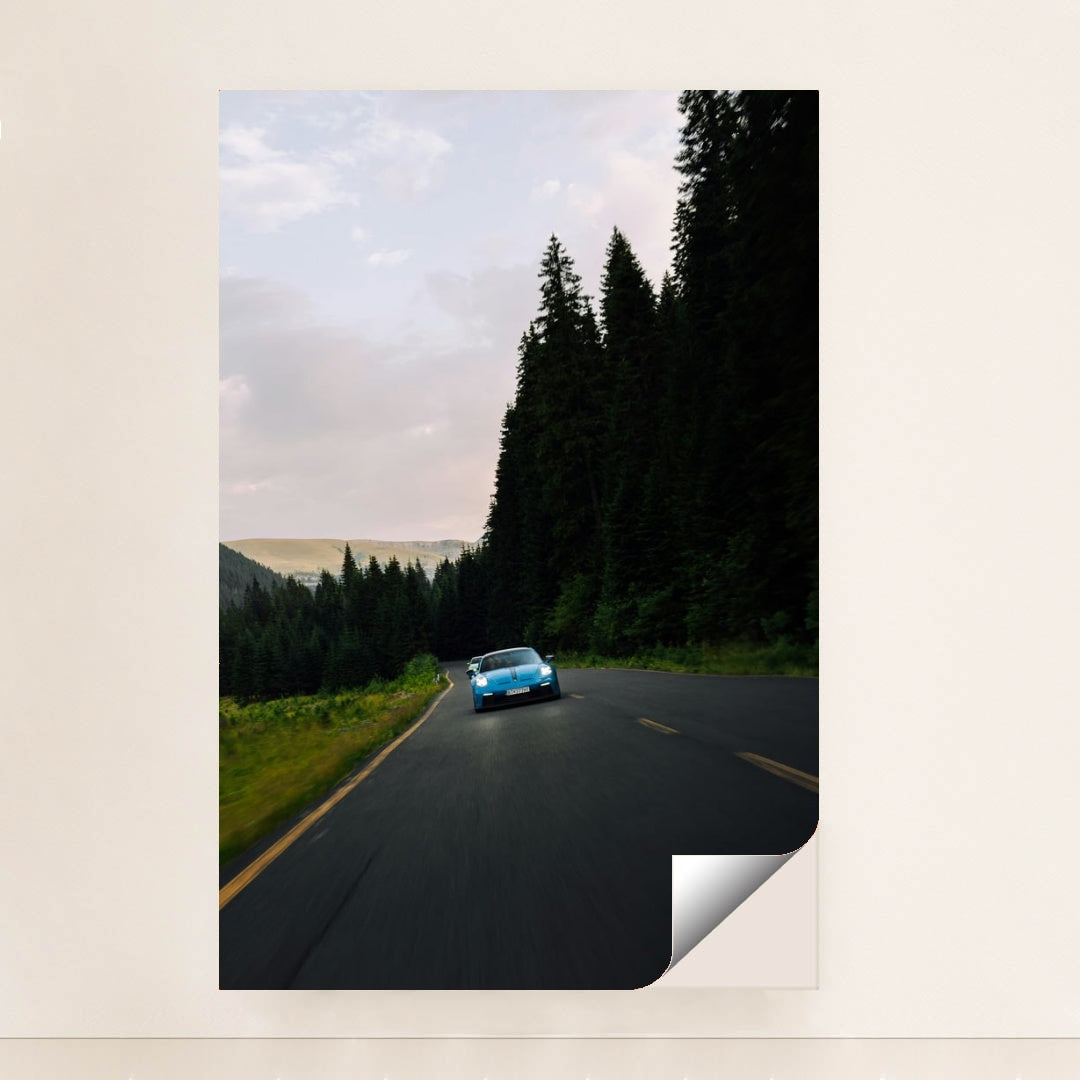 This photo shows a blue Porsche sports car driving on a curvy forest road at dusk, unframed.