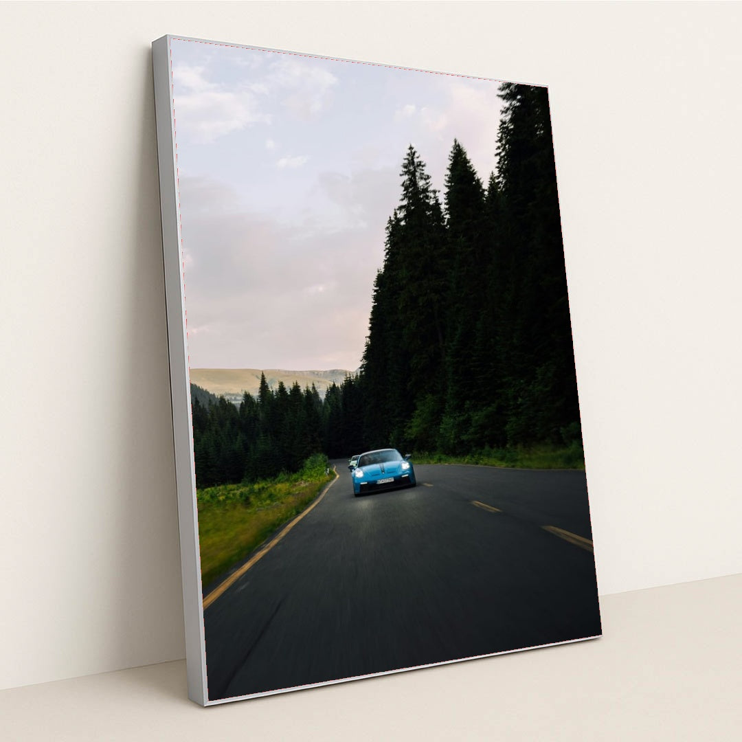 This photo shows a blue Porsche sports car driving on a curvy forest road at dusk, in a silver frame.