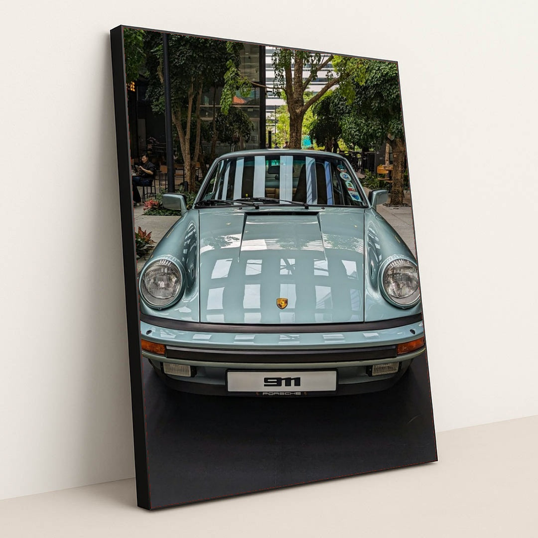 This photo shows a pastel blue classic Porsche 911 coupe on display in a modern indoor courtyard with trees, in a black frame.