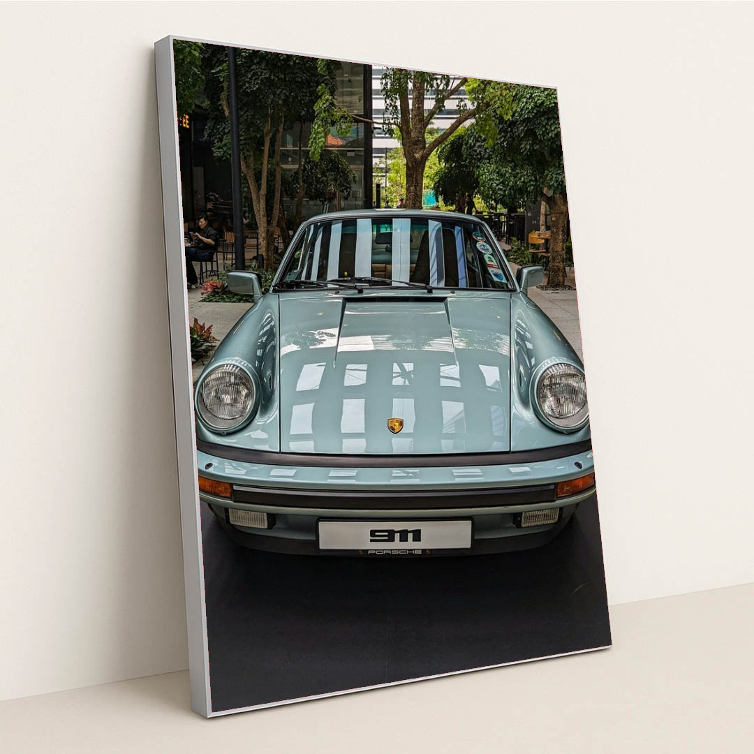 This photo shows a pastel blue classic Porsche 911 coupe on display in a modern indoor courtyard with trees, in a silver frame.