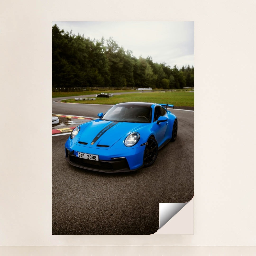 This photo shows a bright blue Porsche 911 GT3 with black racing stripes parked on a curvy racetrack, unframed style.