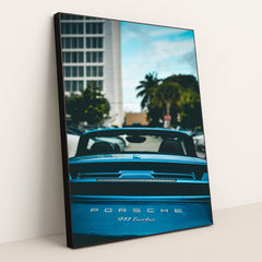 This photo shows the rear view of a blue Porsche 911 Turbo convertible with the top down, parked in a city with palm trees, in a black frame.