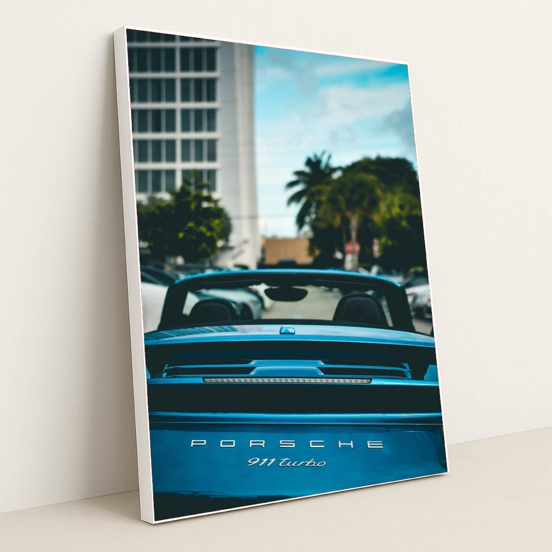 This photo shows the rear view of a blue Porsche 911 Turbo convertible with the top down, parked in a city with palm trees, in a white frame.