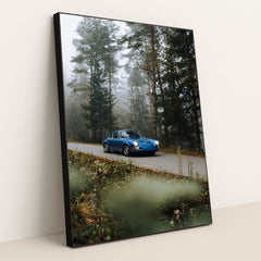 This photo shows a classic blue Porsche driving along a misty forest road with wildflowers in the foreground, in a black frame.
