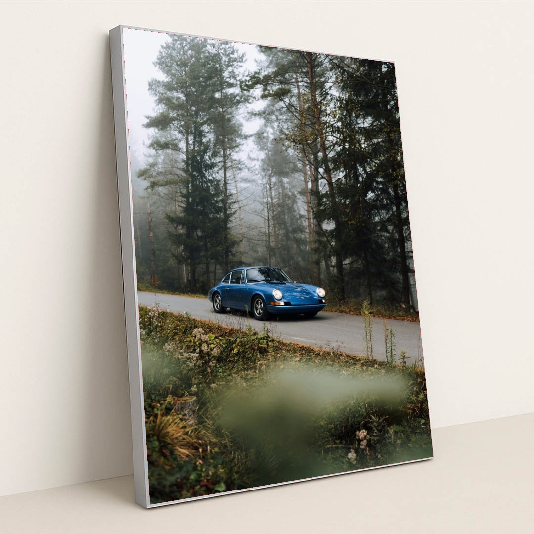 This photo shows a classic blue Porsche driving along a misty forest road with wildflowers in the foreground, in a silver frame.