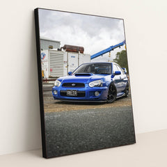 This photo shows a blue Subaru WRX with black wheels parked in an industrial lot, in a black frame.