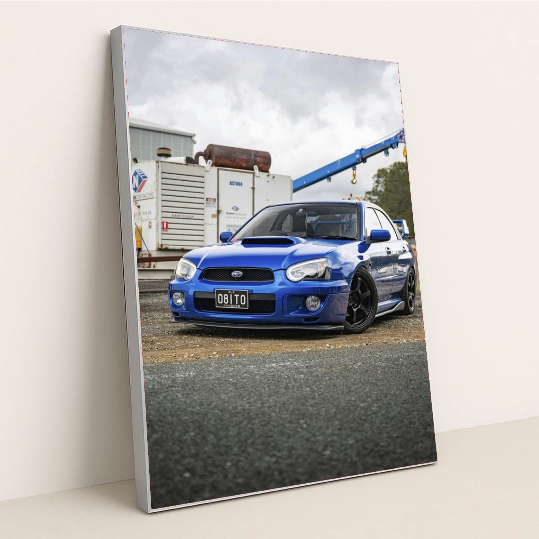 This photo shows a blue Subaru WRX with black wheels parked in an industrial lot, in a silver frame.