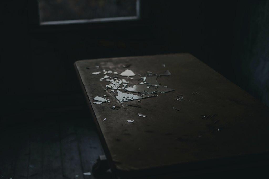 Janko Ferlic - A moody shot of broken glass pieces scattered on an old wooden table in a dimly lit room.