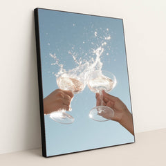 This artwork shows two hands clinking wine glasses with a splash against a clear blue sky, in a black frame.