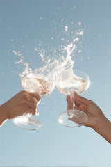 This artwork shows two hands clinking wine glasses with a splash against a clear blue sky.