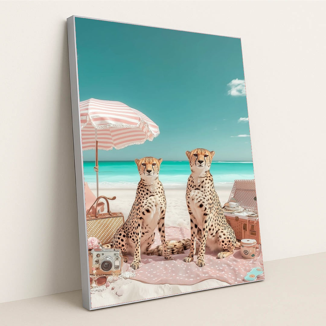This artwork shows two cheetahs sitting on a pink picnic blanket under a striped umbrella at a bright beach, in a silver frame.