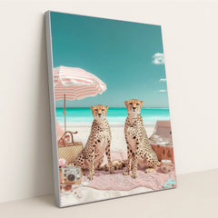 This artwork shows two cheetahs sitting on a pink picnic blanket under a striped umbrella at a bright beach, in a silver frame.