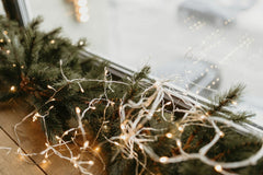 Dmitry Zvolskiy - Close-up of twinkling string lights wrapped around pine garland on a window ledge, creating a festive winter ambiance.
