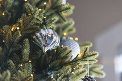 Joshua Wall - Close-up of a Christmas tree with silver ornaments and pinecone, adorned with twinkling lights.