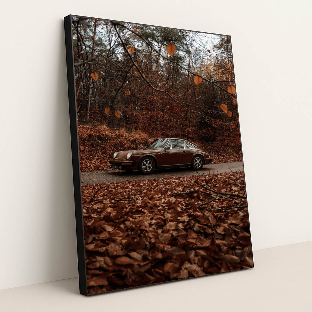 This photo shows a brown classic Porsche 911 coupe parked on a road surrounded by autumn leaves in a forest, in a black frame.