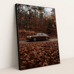 This photo shows a brown classic Porsche 911 coupe parked on a road surrounded by autumn leaves in a forest, in a black frame.