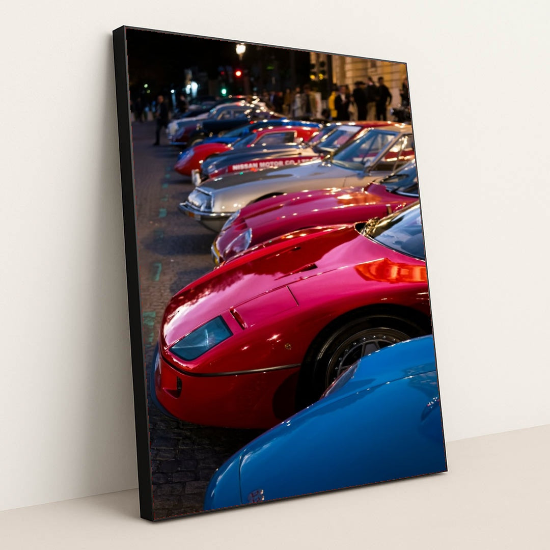 This photo shows a row of vintage sports cars, including a red Ferrari F40, parked on a cobblestone street at dusk, in a black frame.