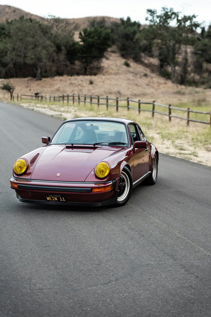 Classic Porsche 911 in California Light parked on a road, featuring yellow headlights and Fuchs wheels, surrounded by hills.