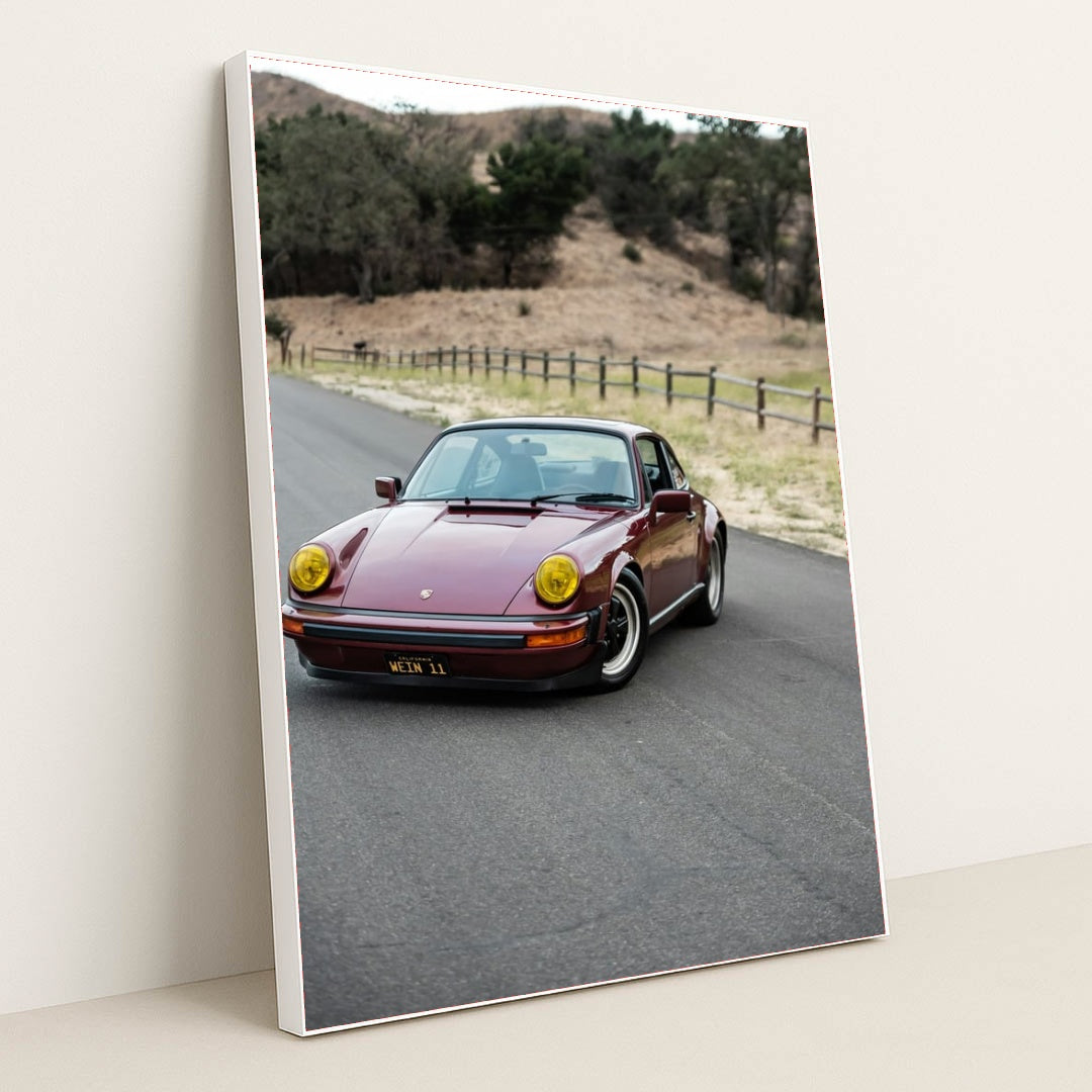 This photo shows a vintage maroon Porsche 911 with yellow headlights driving along a country road, in a white frame.