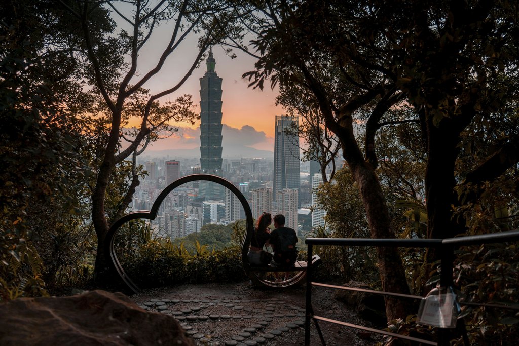 Jimmy Liao - A couple sits on a scenic hill, admiring Taipei 101 at sunset, framed by lush trees.