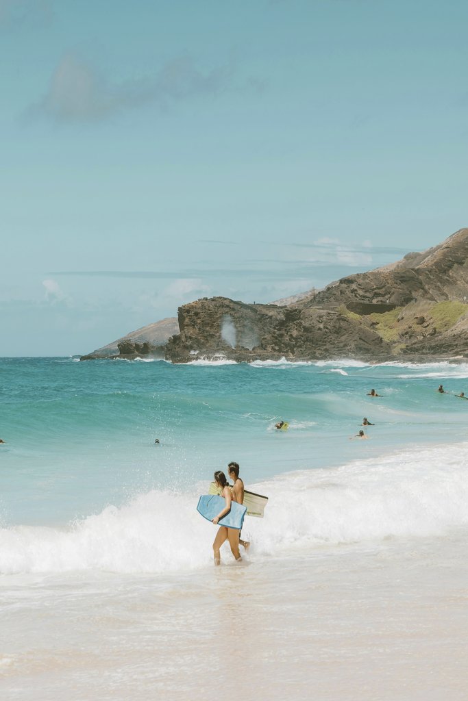 Jess Loiterton - Two surfers with boards walking at a scenic beach with turquoise waves and lush hills.