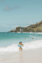 Jess Loiterton - Two surfers with boards walking at a scenic beach with turquoise waves and lush hills.