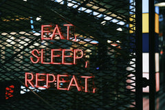 Yusuf Timur Çelik - Close-up of a vibrant neon sign with the slogan 'Eat, Sleep, Repeat' on a metal grate background.