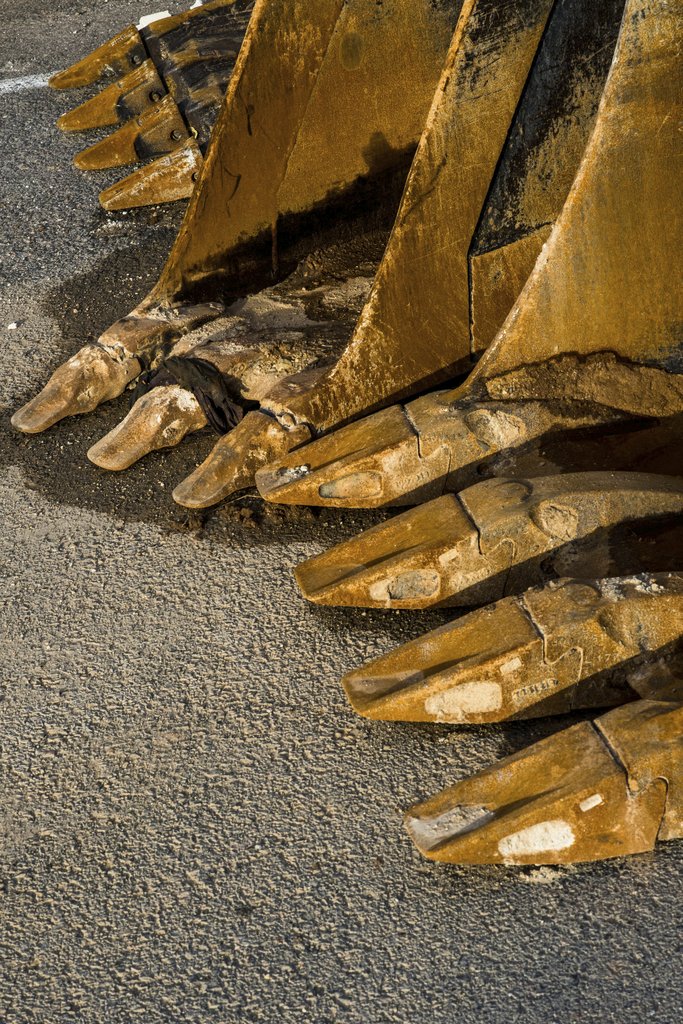 Victor  Moragriega - Detailed view of worn rusted excavator buckets lined up on rough concrete.