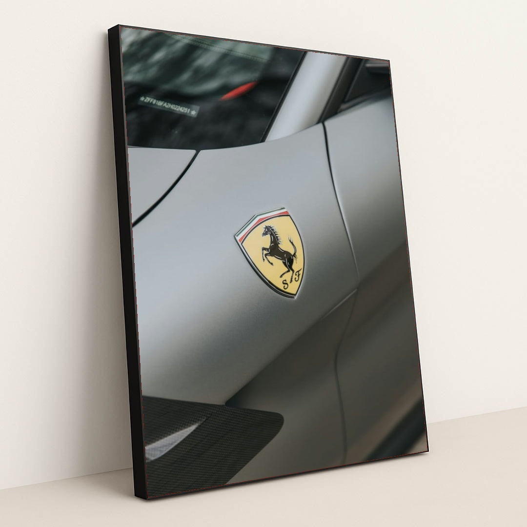 This photo shows a close-up of the Ferrari prancing horse badge on the fender of a matte gray Ferrari, in a black frame.