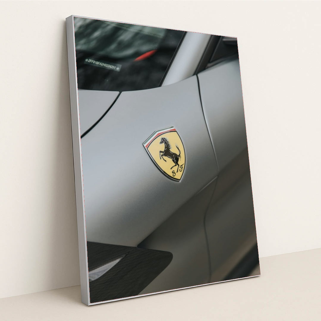 This photo shows a close-up of the Ferrari prancing horse badge on the fender of a matte gray Ferrari, in a silver frame.