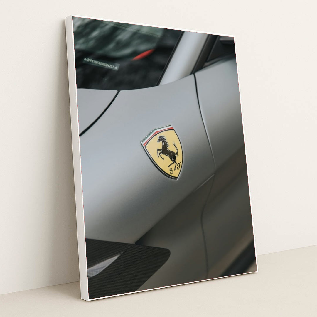 This photo shows a close-up of the Ferrari prancing horse badge on the fender of a matte gray Ferrari, in a white frame.