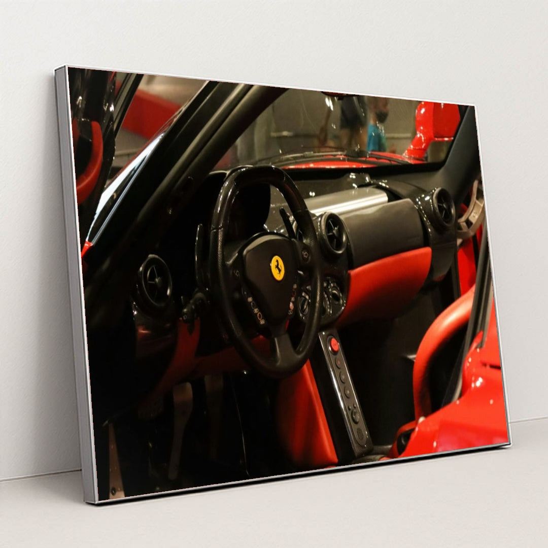 This photo shows the black and red leather interior of a Ferrari, featuring a round steering wheel and minimalist dash, in a silver frame.