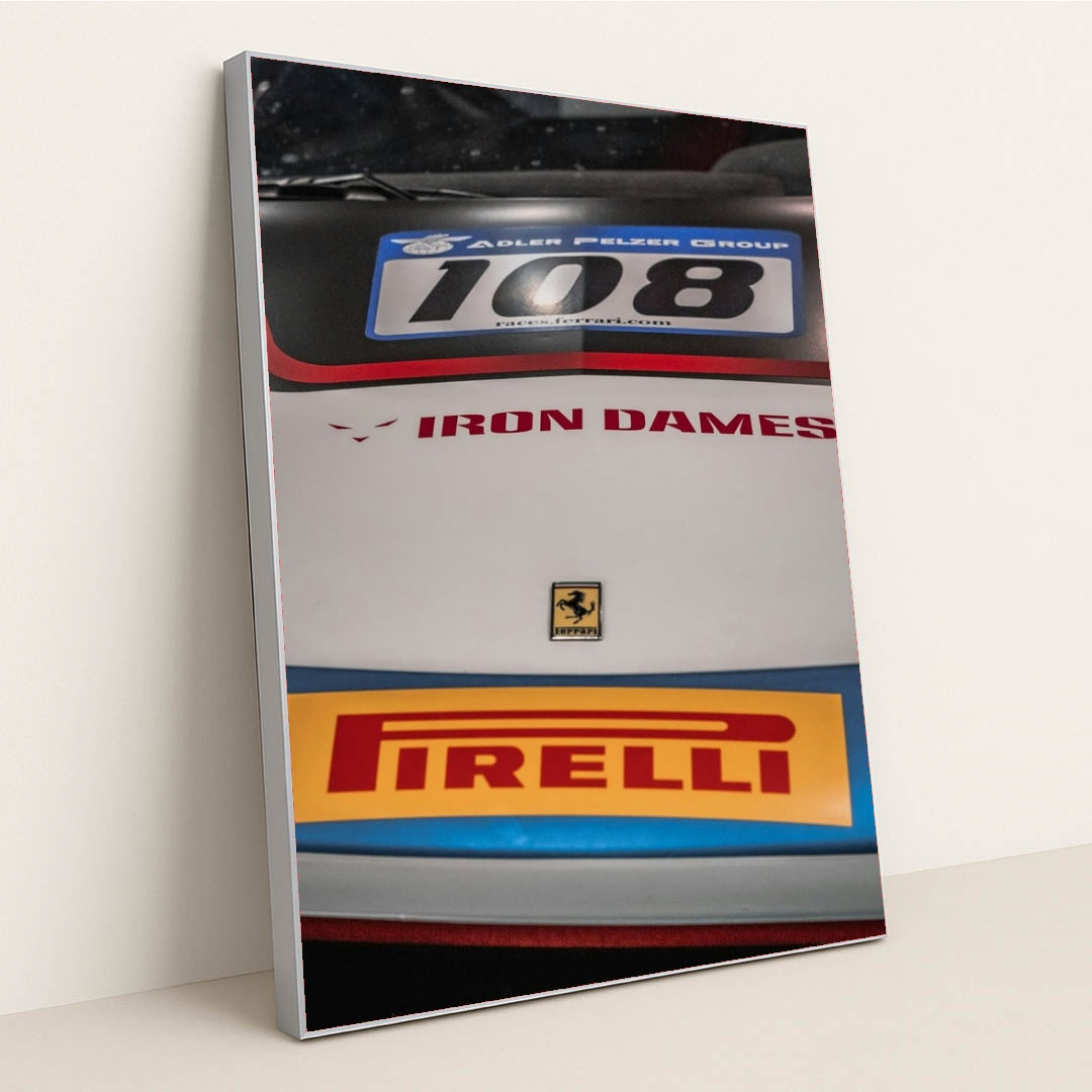 This photo shows a close-up of the hood of a Ferrari race car with Iron Dames and Pirelli logos, in a silver frame.
