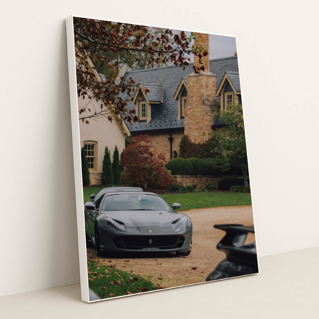 This photo shows a grey Ferrari parked on a stone driveway in front of a large estate with autumn foliage, in a white frame.