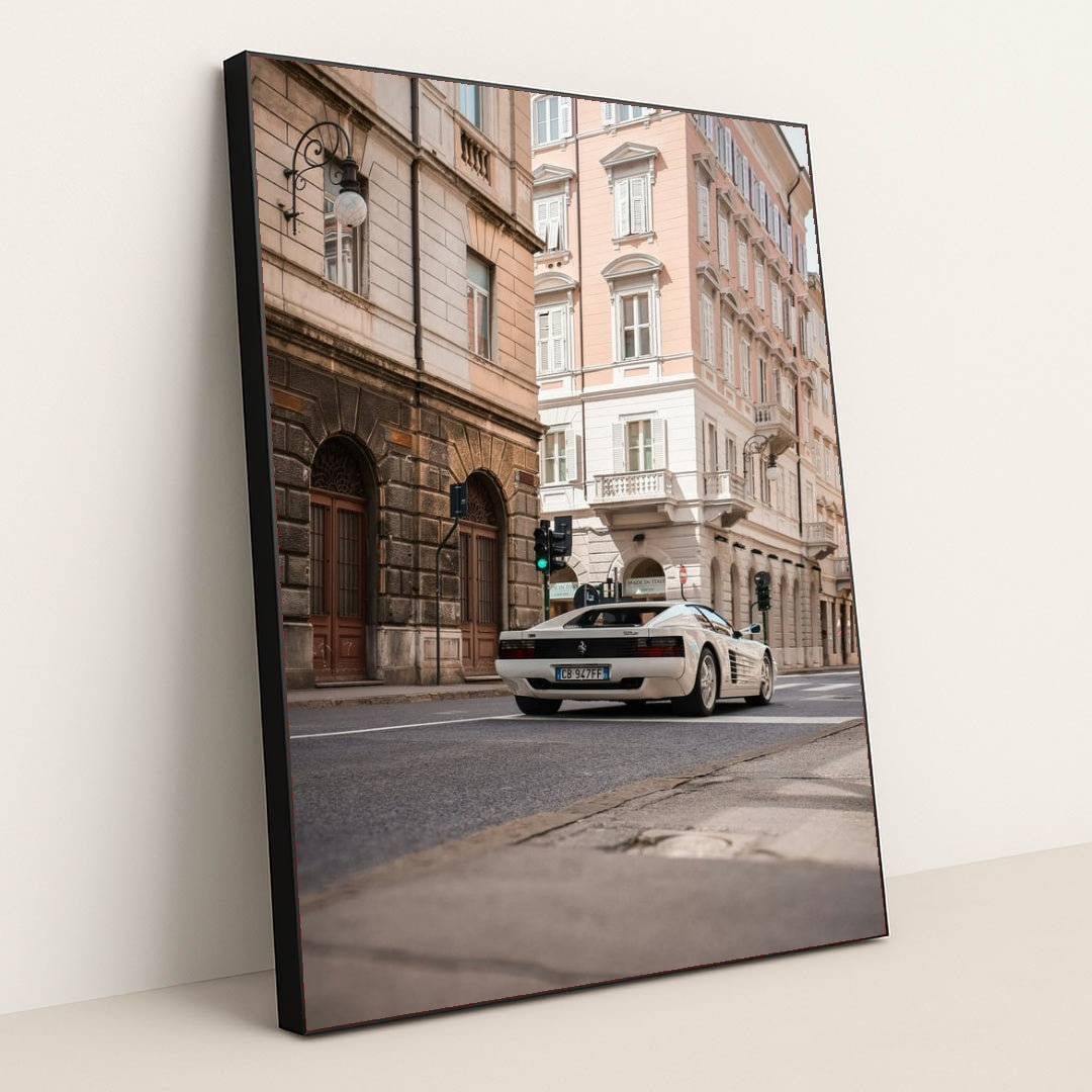 This photo shows a white Ferrari Testarossa driving through a European city street lined with historic pastel buildings, in a black frame.
