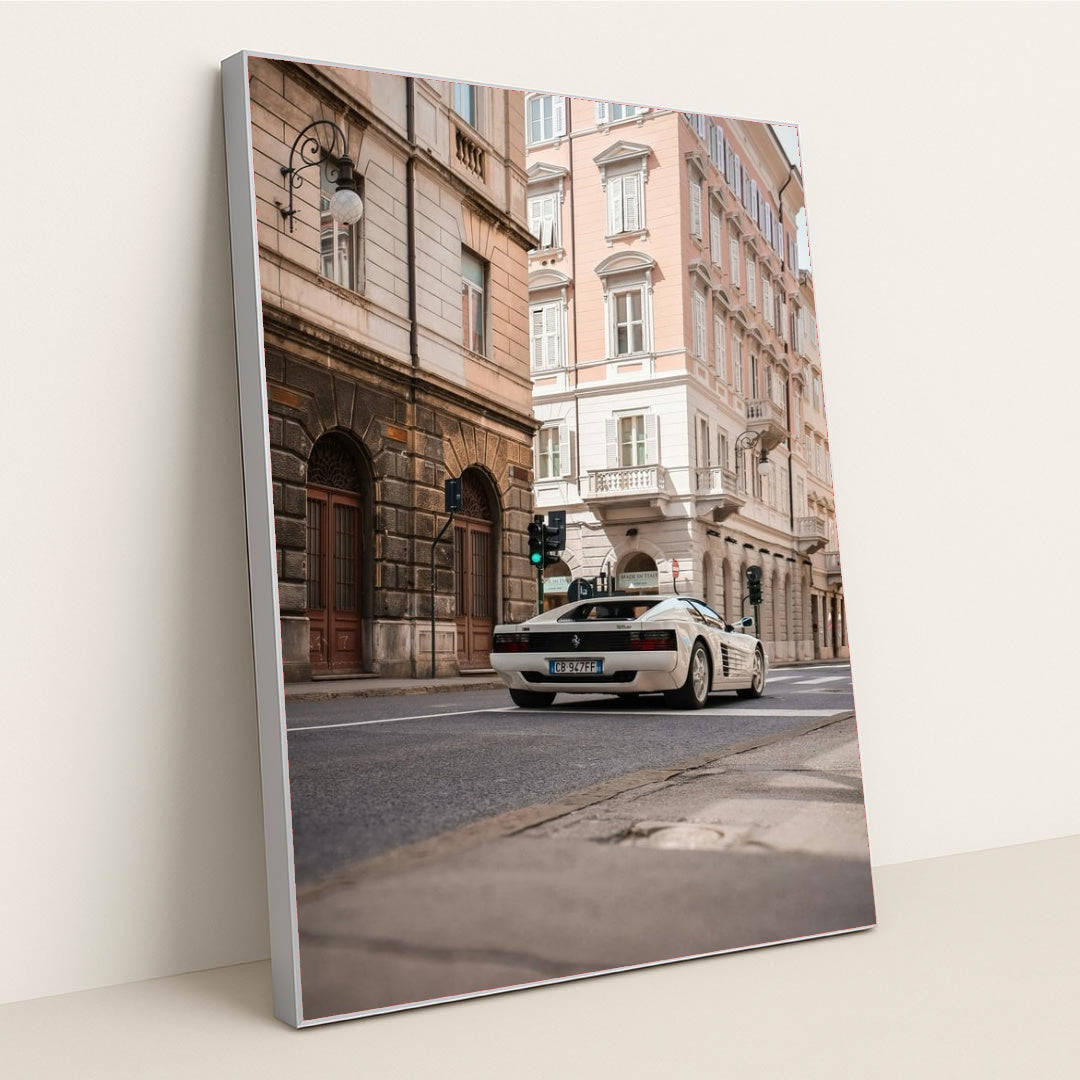 This photo shows a white Ferrari Testarossa driving through a European city street lined with historic pastel buildings, in a silver frame.