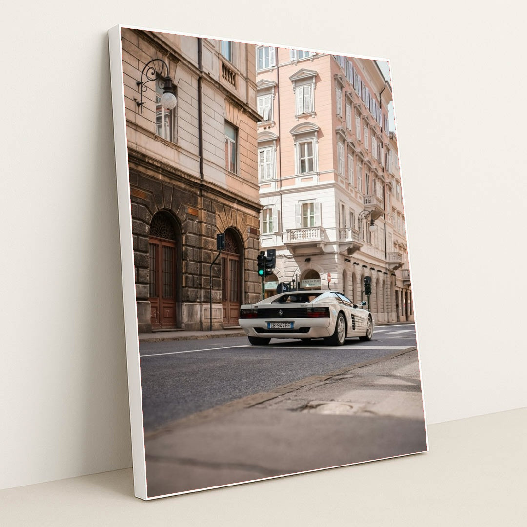 This photo shows a white Ferrari Testarossa driving through a European city street lined with historic pastel buildings, in a white frame.