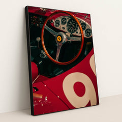 This photo shows the cockpit of a classic red Ferrari race car with a wooden steering wheel and vintage gauges, in a black frame.