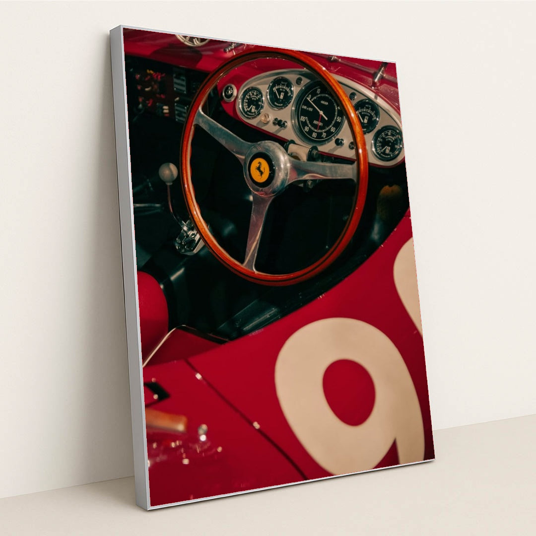 This photo shows the cockpit of a classic red Ferrari race car with a wooden steering wheel and vintage gauges, in a silver frame.